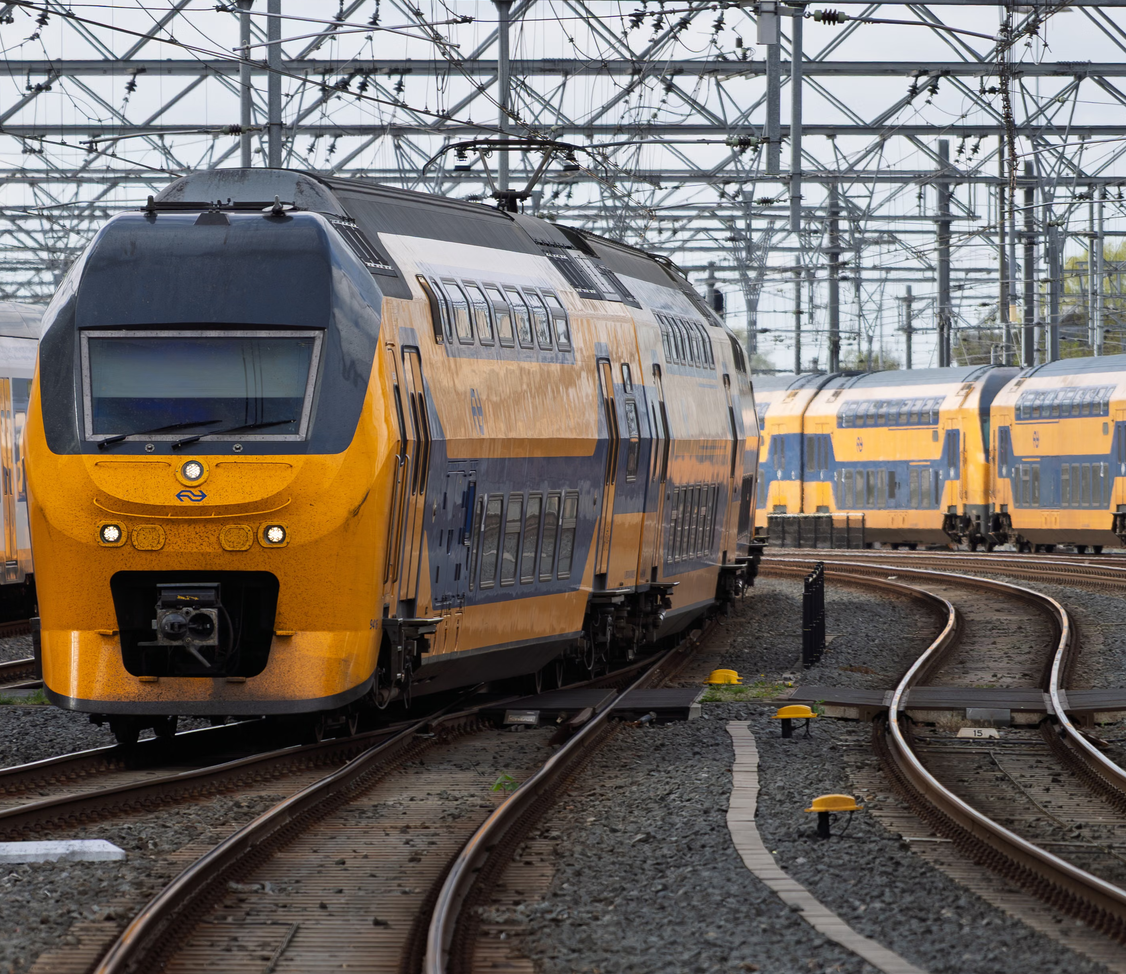 A yellow and blue Dutch train at a railway junction with overhead wires.